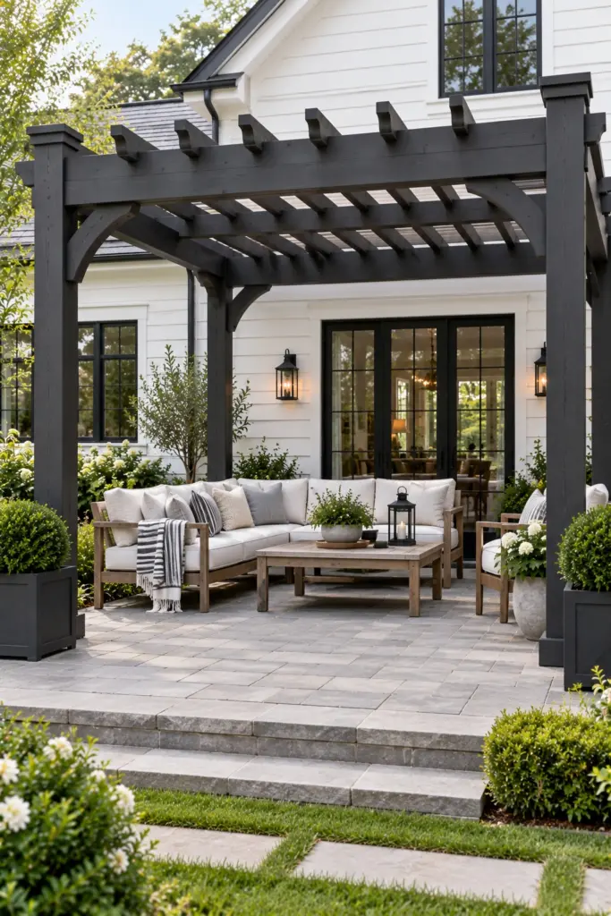 Charcoal solid-stained wooden pergola against white farmhouse siding over light gray pavers